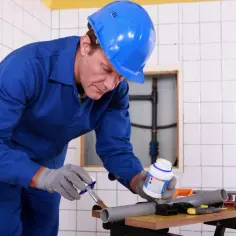 Hydraulik w niebieskim kombinezonie i kasku nakłada klej na rurę PCV, przygotowując ją do połączenia. Pokazuje, jak równo przeciąć rurę pcv.