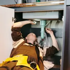 Hydraulik naprawia syfon pod zlewem, gdzie widać zakamienione rury ciepłej wody.