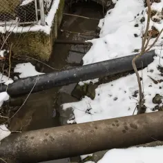 Dwie rury biegną nad strumieniem. Woda w rurach zamarznięta, otoczenie pokryte śniegiem.