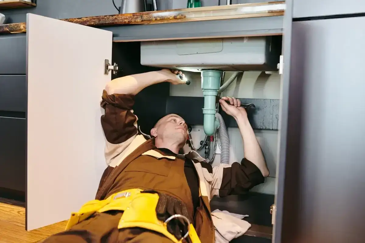 Hydraulik pod zlewem, szukający sposobu, czym rozpuścić kamień w rurach. Używa latarki i klucza.