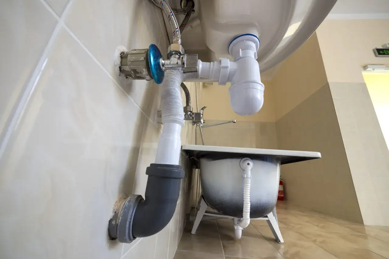 Instalacja hydrauliczna pod umywalką, z widocznym syfonem i odpływem do ściany. To rozwiązanie może pomóc w udrożnieniu rur, jak domowy kret do rur.