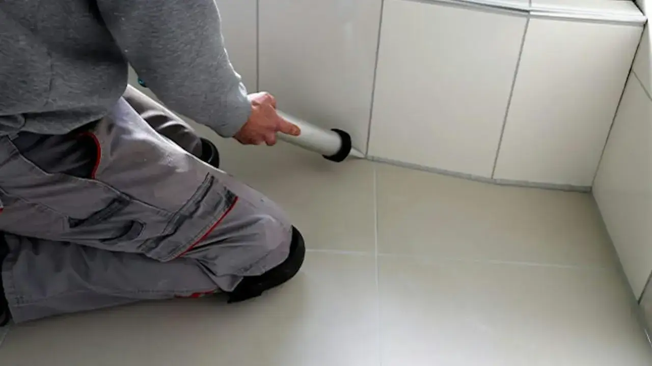 Wykonanie wymiany sylikonu w kabinie prysznicowej. Ręce mężczyzny w szarej bluzie i roboczych spodniach trzymają pistolet do silikonu, aplikując go wzdłuż krawędzi.
